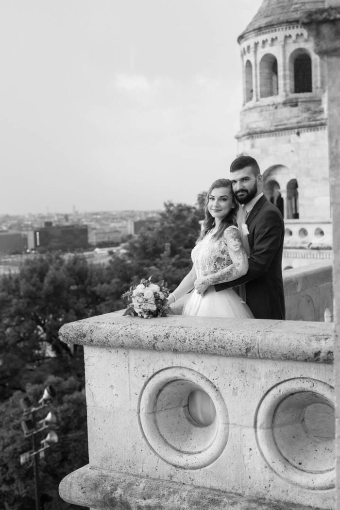 A házaspár romantikus pillanata a Halászbástya falán, miközben a háttérben Budapest városának lenyűgöző panorámája és a történelmi bástya látható. A menyasszony csodálatos ruhájában, a férj elegáns öltönyben, csokorral a kezében, boldog pillanatokban osztozva.