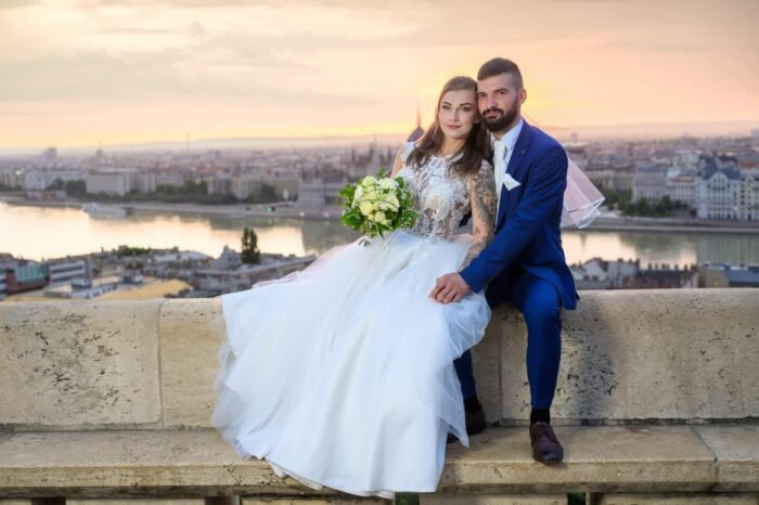 Esküvői fotós Debrecen - A házaspár együtt pihen a Halászbástya falán, a háttérben Budapest lenyűgöző panorámájával és a Duna partján lévő Parlamenttel. A menyasszony csodálatos ruhájában, a férj elegáns öltönyben, csokorral a kezében, boldog pillanatokban osztozva.