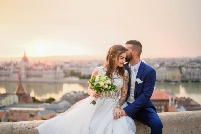 A házaspár együtt pihen a Halászbástya területén, miközben a háttérben a Parlament és a Duna festői panorámája látható. A menyasszony gyönyörű ruhájában, a férj elegáns öltönyben, csokorral a kezében, boldog pillanatokban osztozva.