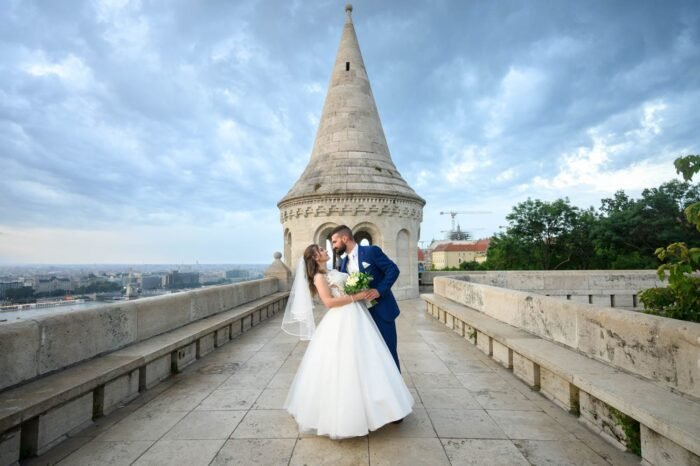A házaspár csókot vált a Halászbástya tetején, a háttérben Budapest lélegzetelállító panorámája és a híres torony látható. A menyasszony gyönyörű ruhájában, a férj elegáns öltönyben, csokorral a kezében.