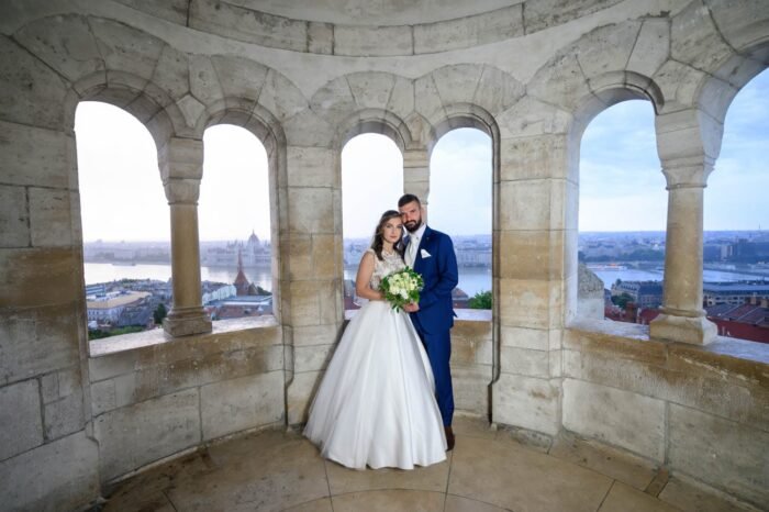 A házaspár boldogan együtt, a Halászbástya belső terében, a háttérben a Duna és a Parlament látványával. A menyasszony csodálatos ruhájában, a férj elegáns öltönyben, csokorral a kezében.