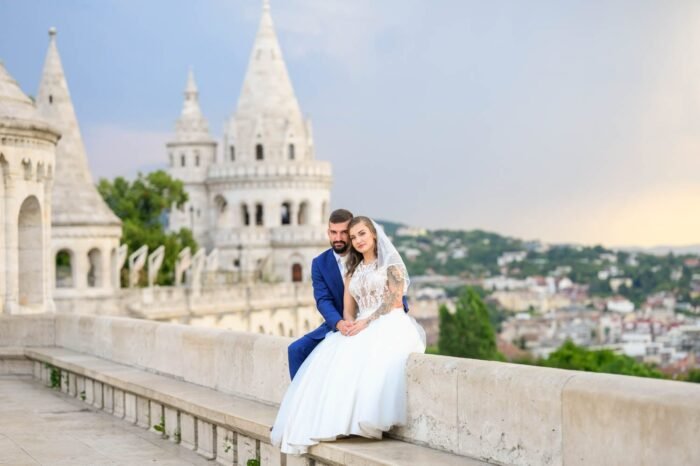 A házaspár együtt pihen a Halászbástya területén, miközben a menyasszony csodálatos ruhájában, a férj elegáns öltönyében van. A háttérben a híres bástya és a város látképe látható, mesés környezetben.