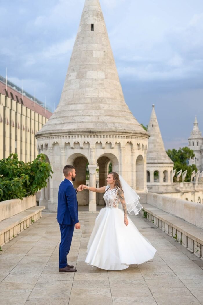 A házaspár együtt táncol a Halászbástya területén, miközben a menyasszony gyönyörű ruhájában, a férj elegáns öltönyében van. A háttérben a híres torony és a bástya egyedülálló építészeti részletei láthatók.