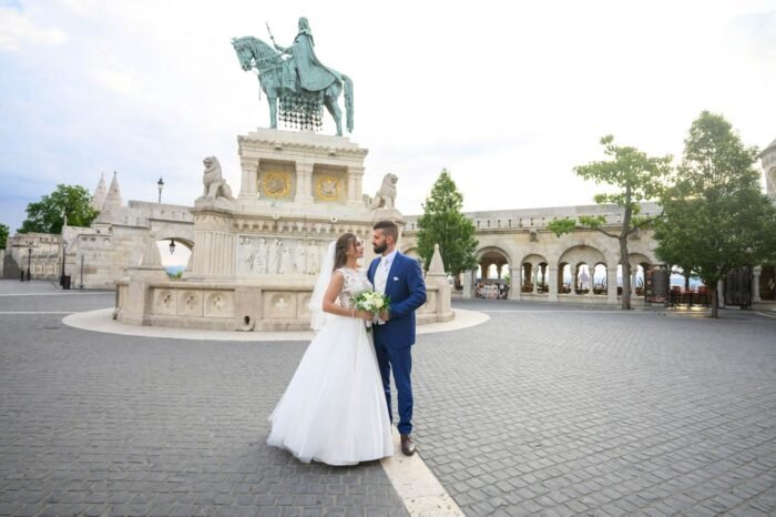 A házaspár boldogan együtt a Halászbástya területén, a háttérben a híres lovas szoborral és a történelmi építészeti részletekkel. A menyasszony csodálatos ruhájában, a férj elegáns öltönyben, csokorral a kezében.