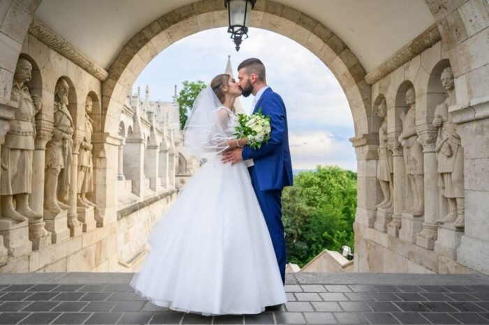 A friss házas pár romantikus csókot vált a Halászbástya híres ívénél, a háttérben a történelmi építészeti részletekkel és a zöld tájjal. A menyasszony csipkés ruhájában, a férj elegáns öltönyben, csokorral a kezében.