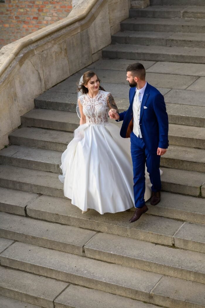 A házaspár kézen fogva sétál a Halászbástya lépcsőin, miközben a menyasszony gyönyörű, csipkés ruhájában, a férj elegáns öltönyében van. A háttérben a történelmi épület és a lépcsők láthatók, a pár boldog pillanatai.