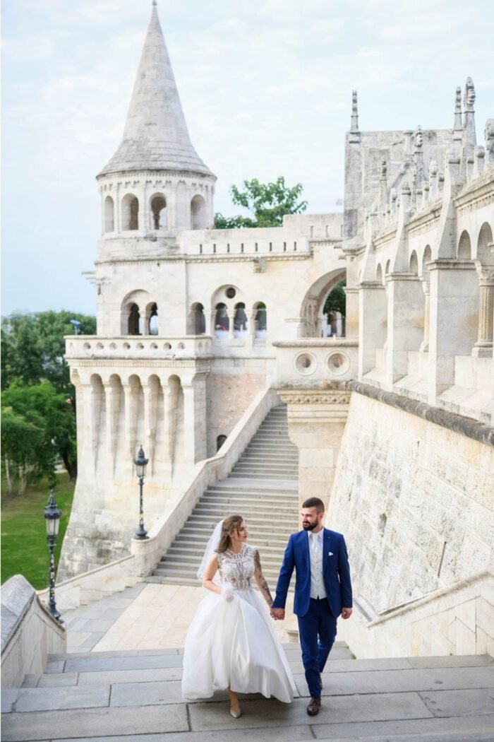 A házaspár kézen fogva sétál a Halászbástya lépcsőin, miközben a menyasszony gyönyörű, csipkés ruhájában és a férj elegáns öltönyében van. A háttérben a mesés építészeti részletek és a zöld táj látható.