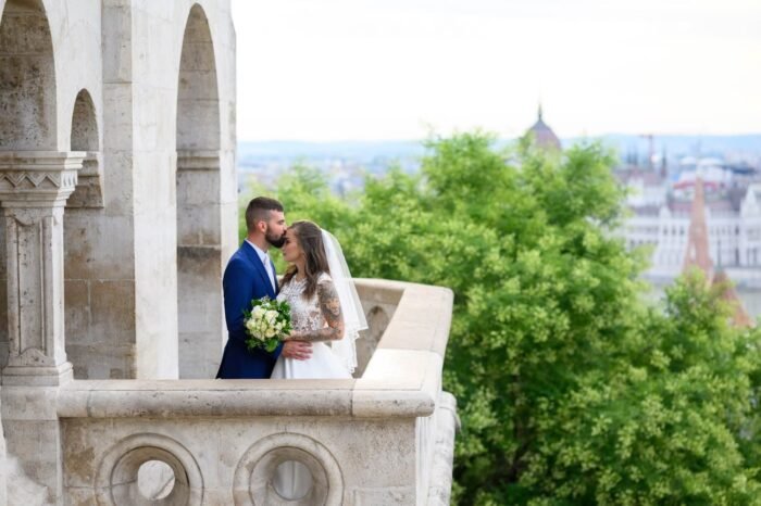 A boldog házaspár csókot vált a Halászbástya kilátóján, miközben a háttérben Budapest lenyűgöző panorámája látszik. A menyasszony elegáns ruhában, a férj öltönyben, csokorral a kezében.