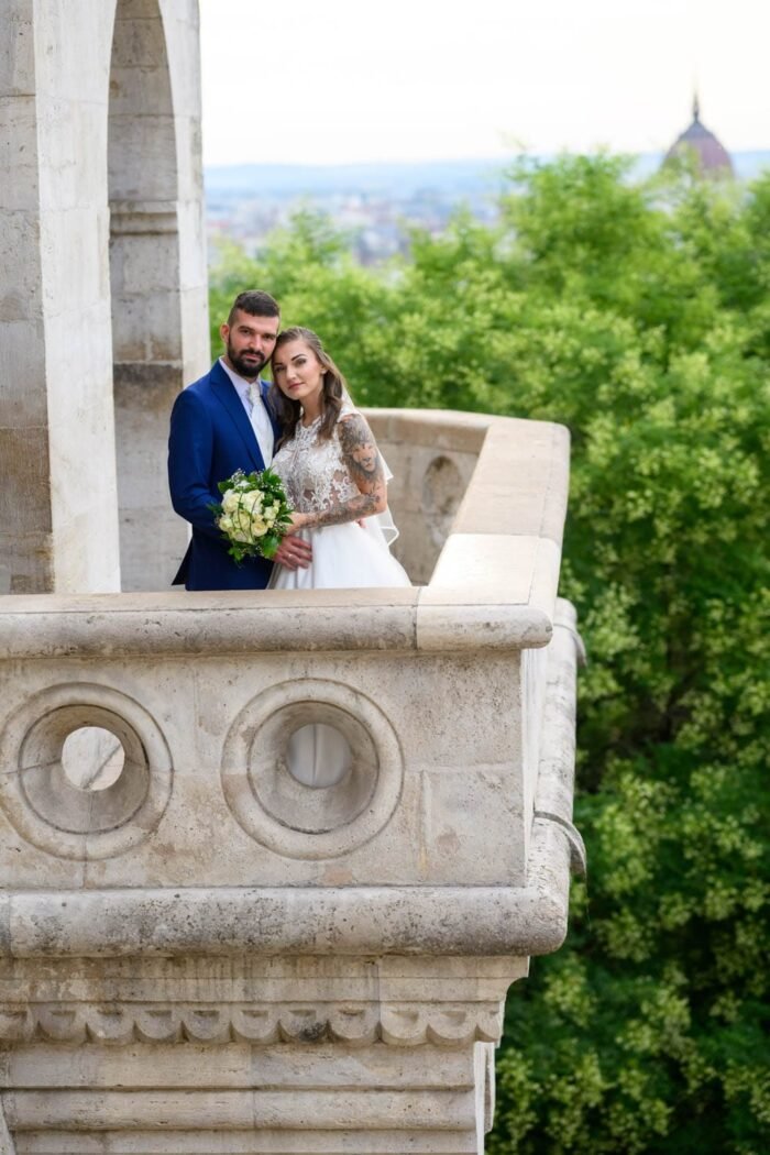 A friss házas pár boldogan együtt, a Halászbástya kilátóján, a menyasszony gyönyörű ruhájában, a férj elegáns öltönyben, miközben Budapest festői panorámája és zöld tája adja a háttért. A pár közel áll egymáshoz, kezük összeér, miközben a menyasszony csokrot tart.