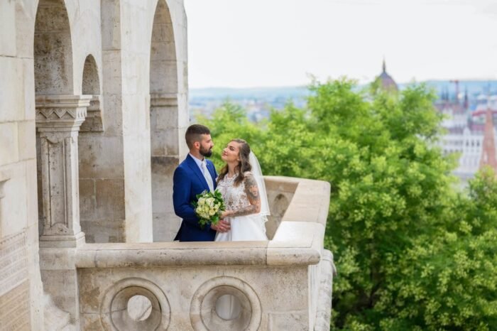 A házaspár boldog pillanata a Halászbástya kilátóján, a menyasszony csodálatos ruhájában, a férj elegáns öltönyben, miközben Budapest lenyűgöző panorámája és zöld tája adja a háttért. A pár közel áll egymáshoz, miközben a menyasszony csokrot tart a kezében.