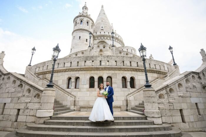 A friss házas pár együtt áll a Halászbástya lépcsőjén, a menyasszony csodálatos ruhájában, a férj elegáns öltönyben, miközben a háttérben Budapest történelmi építészete és a híres műemlékek láthatók.