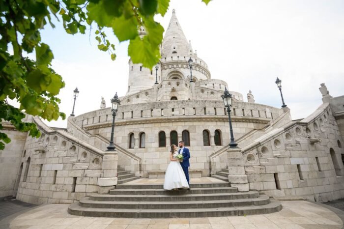 A friss házas pár romantikus pillanata a Halászbástya lépcsőjén, háttérben a híres építészeti műemlék és a történelmi Budapest látképével. A menyasszony elegáns fehér ruhában, a férj öltönyben, csokorral a kezében.