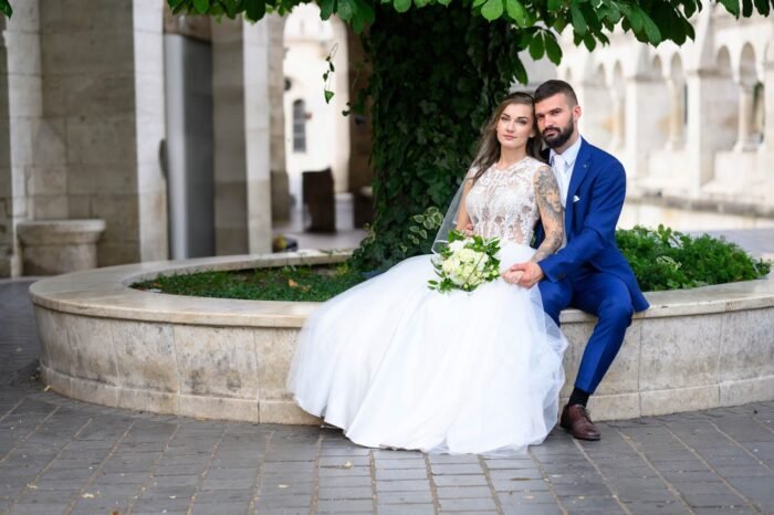 Kreatív fotózás Budapesten. A házaspár együtt pihen a parkban, a menyasszony elegáns, csipkés ruhájában, míg a férj elegáns öltönyben ül mellette, kezük összeér, a háttérben történelmi épület és zöld növényzet látható.