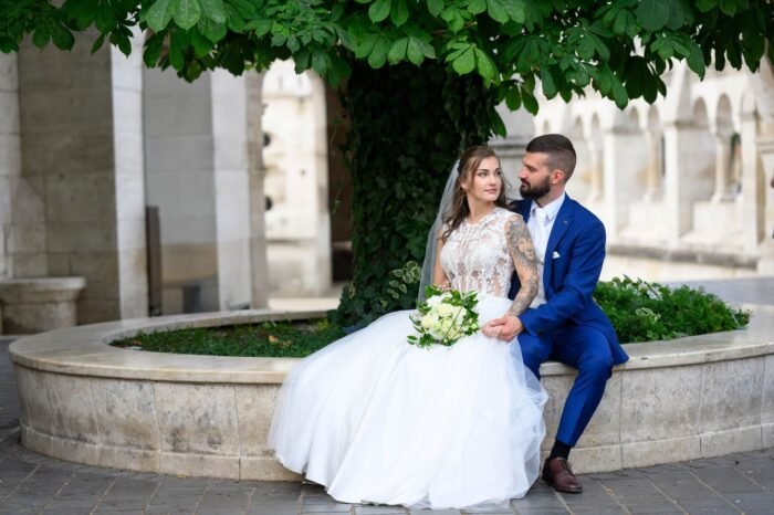 A friss házas pár egy parkban pihen, menyasszonyi ruhában és öltönyben, miközben egymásra néznek, a háttérben a történelmi építészet és zöld növényzet díszíti a jelenetet.