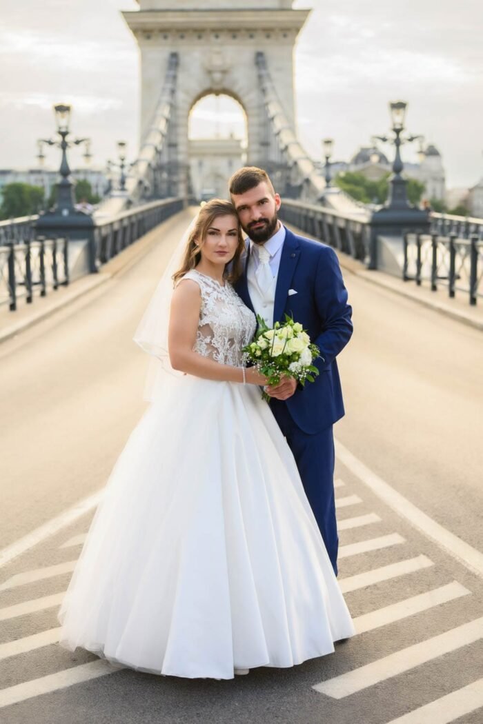 Esküvői fotózás Budapesten. Friss házasok romantikus pillanata Budapest ikonikus Lánchídján, a fiatal pár menyasszonyi ruhában és öltönyben, csokorral a kezükben.