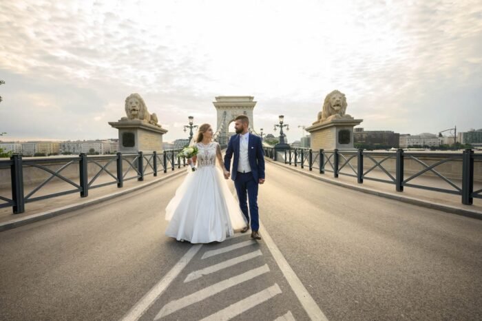 Esküvői pár kézenfogva sétál a Lánchídon, háttérben a híres oroszlánszobrok és Budapest városképe. A menyasszony fehér, elegáns ruhában, a vőlegény stílusos kék öltönyben látható, miközben a reggeli fény lágyan megvilágítja őket.
