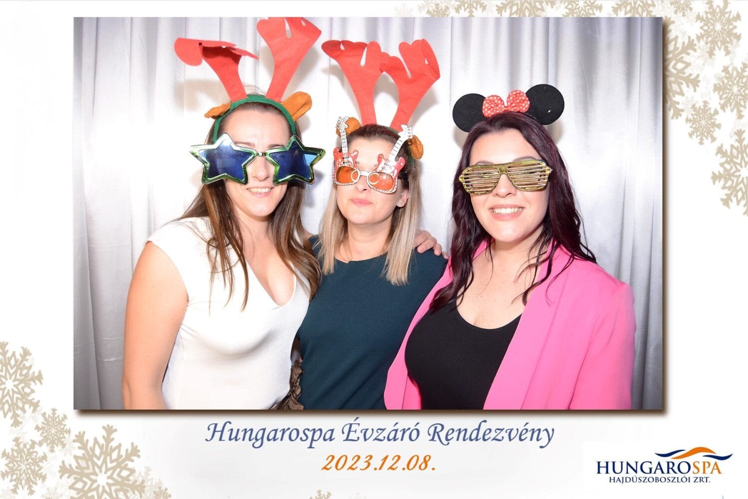 Három mosolygós nő jelmezekben és vicces kiegészítőkkel pózol egy fotóbox előtt a Hungarospa Évzáró Rendezvényén, 2023. december 8-án. A kép alján rendezvény információk és a Hungarospa logója látható.