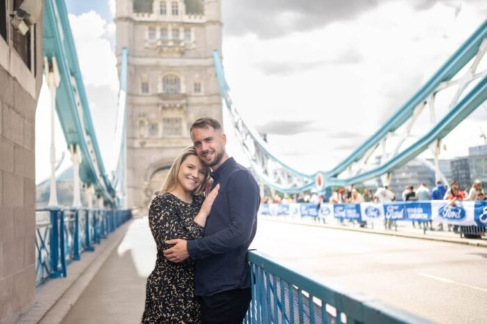 Egy mosolygó jegyespár áll a londoni Tower Bridge-en, szorosan egymáshoz bújva, a híres híd ikonikus kék acélszerkezete és tornyai előtt, amely különleges hátteret ad a romantikus pillanatnak.