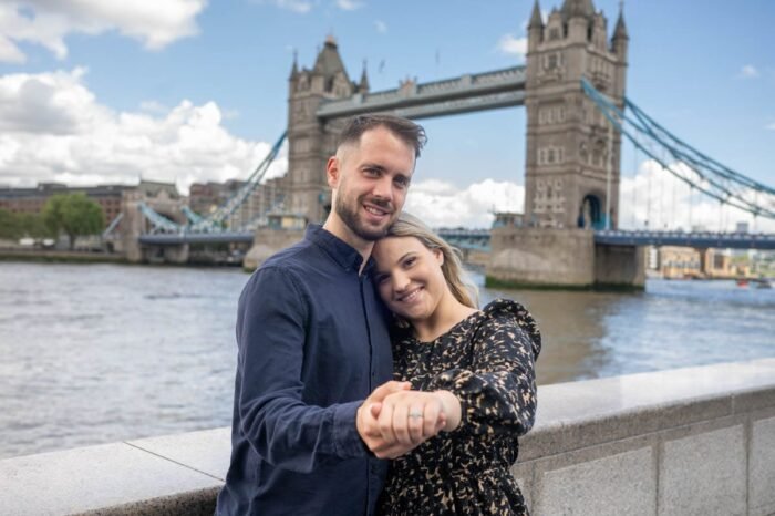 Egy mosolygó jegyespár Londonban, a Tower Bridge ikonikus hátterében, a lány ujján frissen felhúzott eljegyzési gyűrűvel, romantikus pillanatot megörökítve.