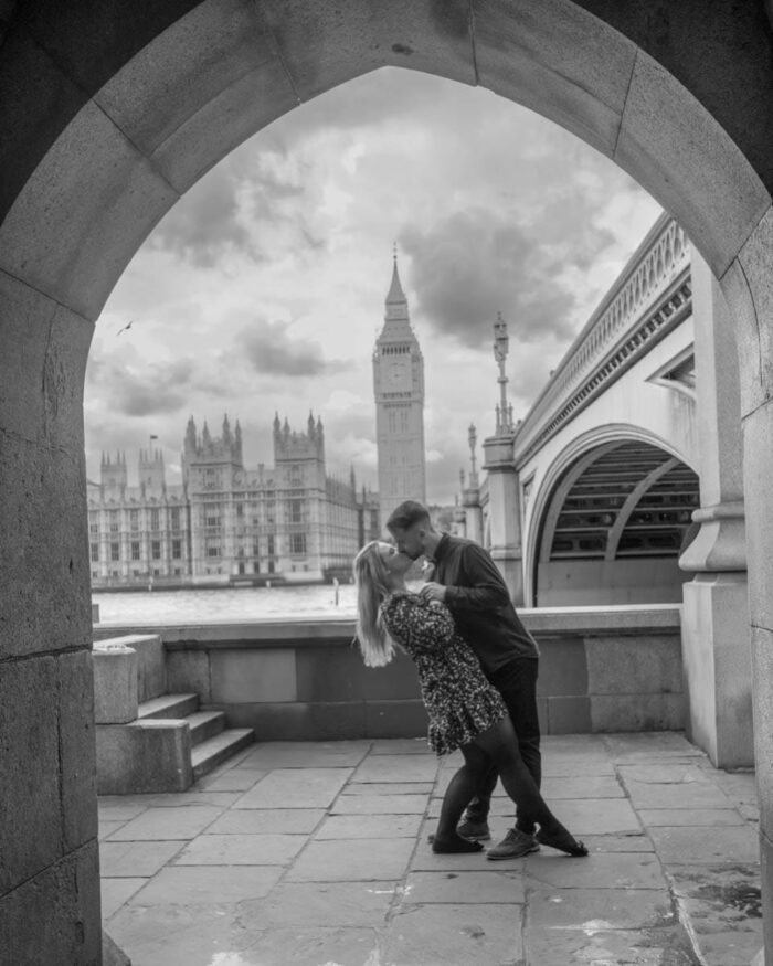 Egy jegyespár romantikus csókot vált Londonban, a Big Ben és a Westminster híd ikonikus látványával a háttérben, egy boltív alól nézve.