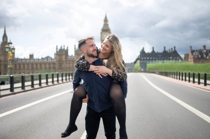 Egy fiatal pár a Westminster hídon, háttérben a Big Ben és a Parlament épülete. A férfi hátán viszi a nőt, mindketten nevetnek. A nő fekete-arany mintás ruhát és fekete harisnyát, a férfi sötétkék inget és fekete nadrágot visel.