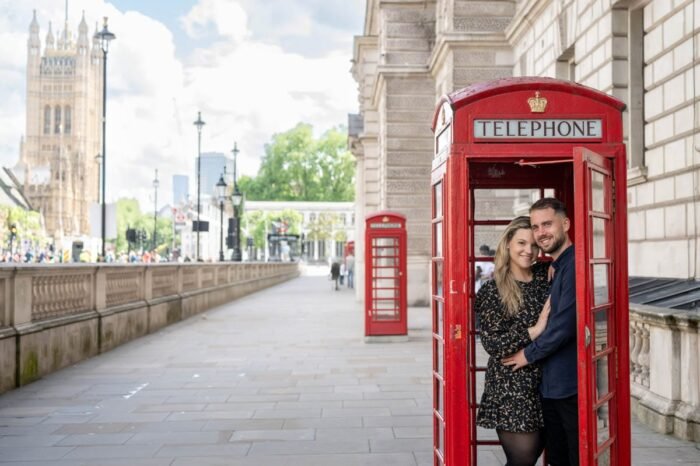 Jegyespár egy piros londoni telefonfülkében mosolyog, háttérben a Westminster-palota tornyával. A nő fekete-arany mintás ruhát, a férfi sötétkék inget visel. A képen jól látható London klasszikus utcaképe a viktoriánus lámpákkal és kőépületekkel.
