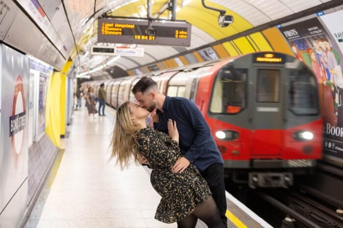Fiatal pár csókolózik egy londoni metróállomáson, a háttérben egy piros metrószerelvény látható. A nő fekete-arany mintás ruhát visel, a férfi sötétkék felsőt, romantikus mozdulattal hajolnak egymáshoz.