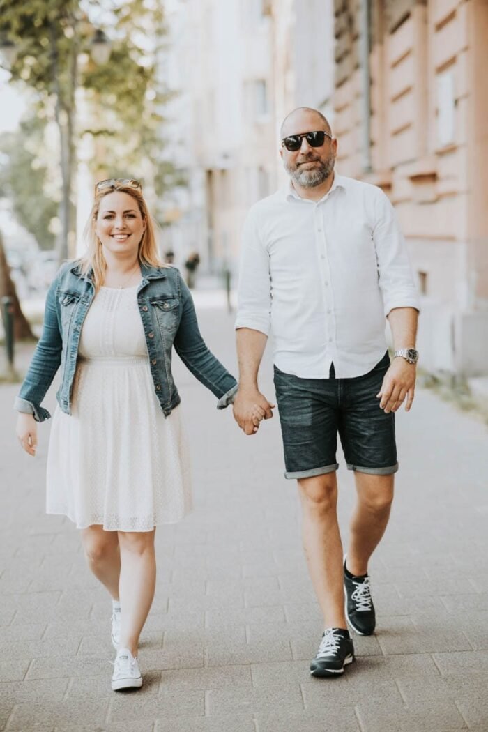 A pár kézen fogva sétál egy városi utcán, boldogan mosolyognak. A nő fehér ruhát visel, amelyet egy farmerdzseki egészít ki, a férfi pedig fehér inget és rövidnadrágot visel. Mindketten fehér sportcipőt viselnek, és napszemüveget hordanak. A háttérben városi épületek és fák láthatók, ami egy nyári, napfényes hangulatot sugároz.