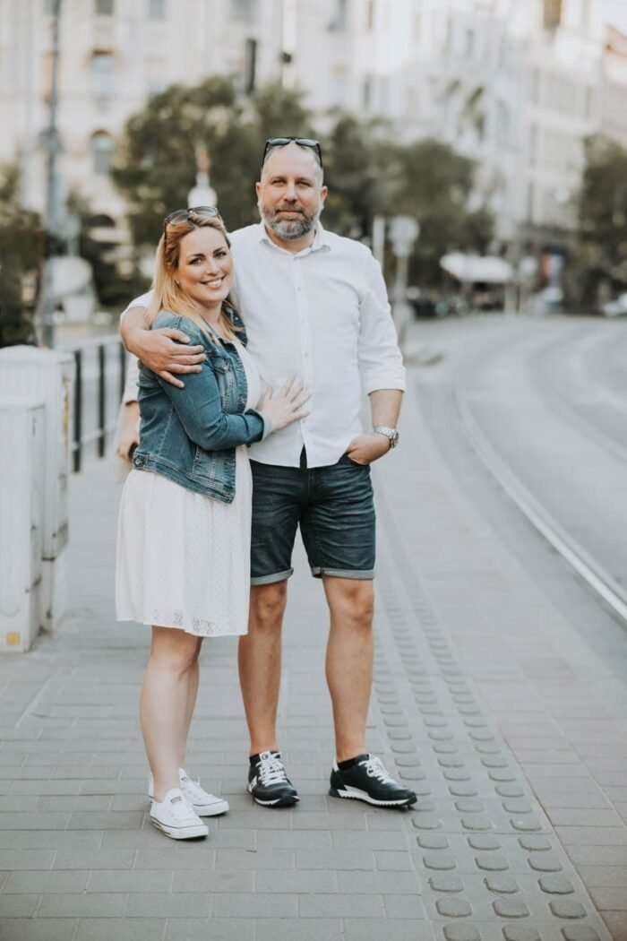 Egy pár sétál a város egyik utcáján, kezüket összefonva. A nő fehér ruhát visel, amit egy farmerdzseki egészít ki, míg a férfi fehér inget és rövidnadrágot visel. Mindketten sportcipőt hordanak. A háttérben a város épületei és egy villamos sín látható, ami az utcán halad. A pár boldogan mosolyog egymásra, miközben az utca csendes, nyugodt hangulatot sugároz.