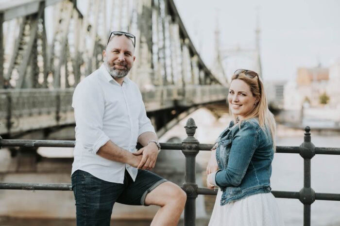 Egy pár boldogan pózol a híddal a háttérben. A férfi fehér inget visel és rövidnadrágot, míg a nő fehér ruhát és farmerdzsekit visel. Mindketten napszemüveget hordanak, és a férfi egyik kezével a korláton pihen, míg a nő boldogan mosolyogva néz a fényképezőgép felé. A háttérben látható a híd, amely szimbolizálja a város egyik nevezetességét.