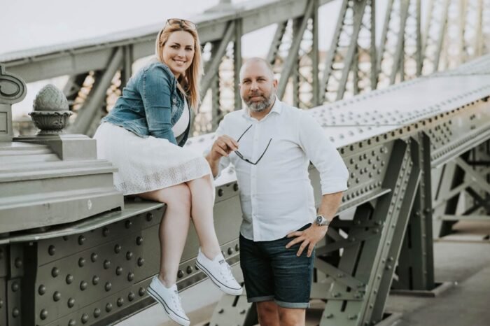 Egy pár romantikus jegyes fotózáson vesz részt Budapest híres hídján. A nő fehér ruhát visel, farmerdzsekivel és fehér sportcipővel, miközben a híd szélére ült, a férfi pedig mellette áll, fehér ingben és rövidnadrágban. A háttérben jól látszik a híd szerkezete, és a város nyújtja a festői hátteret. Mindketten boldogan mosolyognak a kamerába, miközben a férfi napszemüveget tart a kezében.