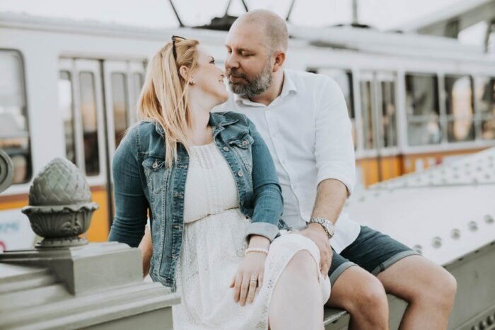 Egy pár romantikus pillanatokat oszt meg egymással a budapesti hídon, miközben a nő ül a híd szélén, és a férfi közel áll hozzá. A háttérben egy villamos halad el, ami Budapest jellegzetes közlekedési eszköze. Mindketten boldogan mosolyognak egymásra, miközben gyengéden néznek egymás szemébe, a férfi napszemüveget tart a kezében. A nő fehér ruhát visel, farmerdzsekivel, és fehér sportcipőt, míg a férfi fehér inget és rövidnadrágot visel.