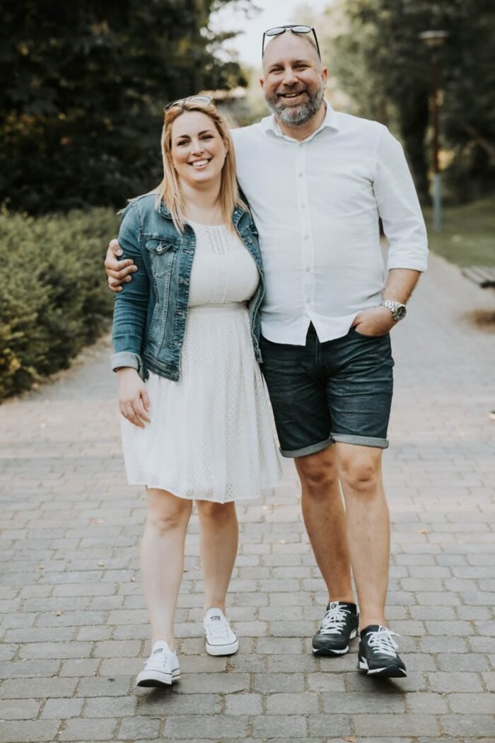 Egy pár boldogan sétál egy parkban, miközben kézen fogva mosolyognak egymásra. A nő fehér ruhát visel, farmerdzsekivel és fehér sportcipővel, míg a férfi fehér inget és rövidnadrágot visel. Mindketten kényelmesen öltözködtek, és jókedvűen élvezik a sétát. A háttérben egy zöld terület látható, amit a napsütés világít meg.