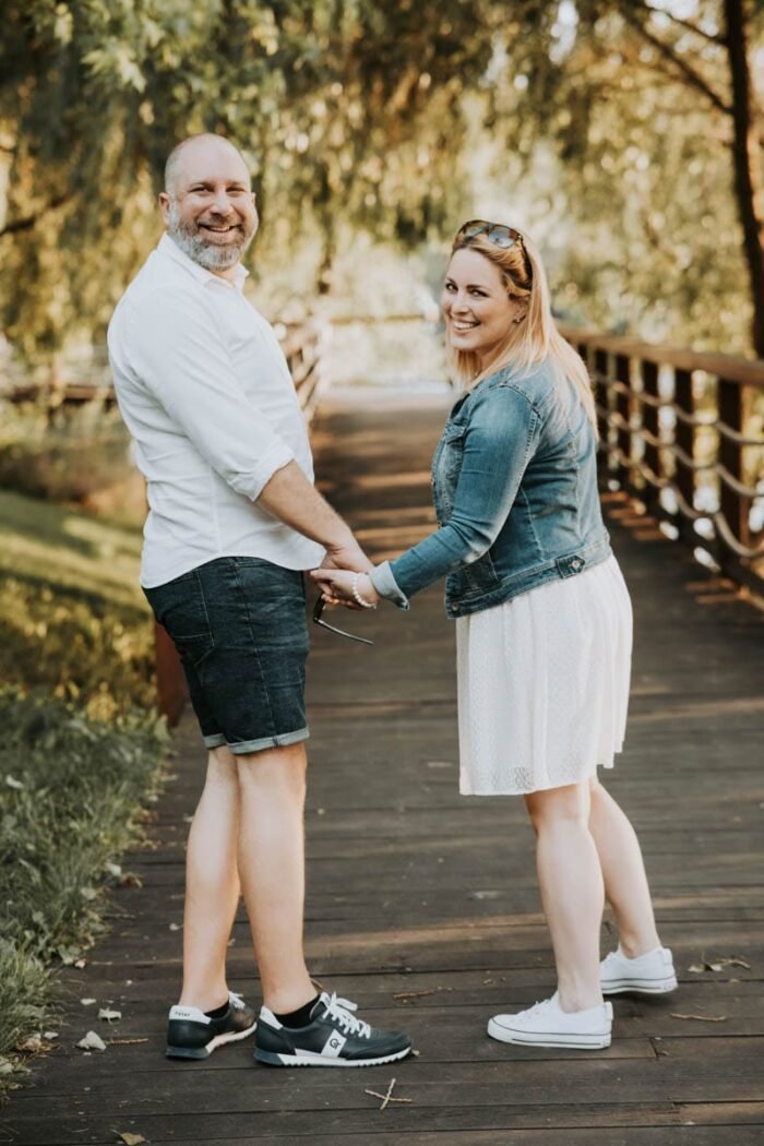 A pár boldogan sétál a hídon, miközben kézen fogva mosolyognak egymásra. A férfi fehér inget és rövidnadrágot visel, míg a nő fehér ruhát és farmerdzsekit visel. Mindketten kényelmes sportcipőt viselnek, és egy napsütéses, zöld parkos környezetben járnak. A háttérben egy fa híd és a természet szépsége látható, amit a meleg fények világítanak meg.