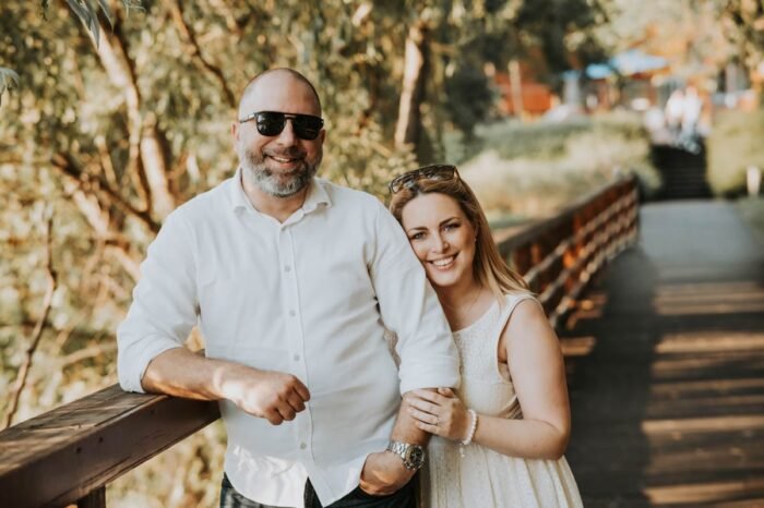 Mosolygós pár áll egy fából készült híd korlátjánál, napsütötte természeti környezetben. A férfi fehér ingben és napszemüvegben, míg a nő világos ruhában látható.