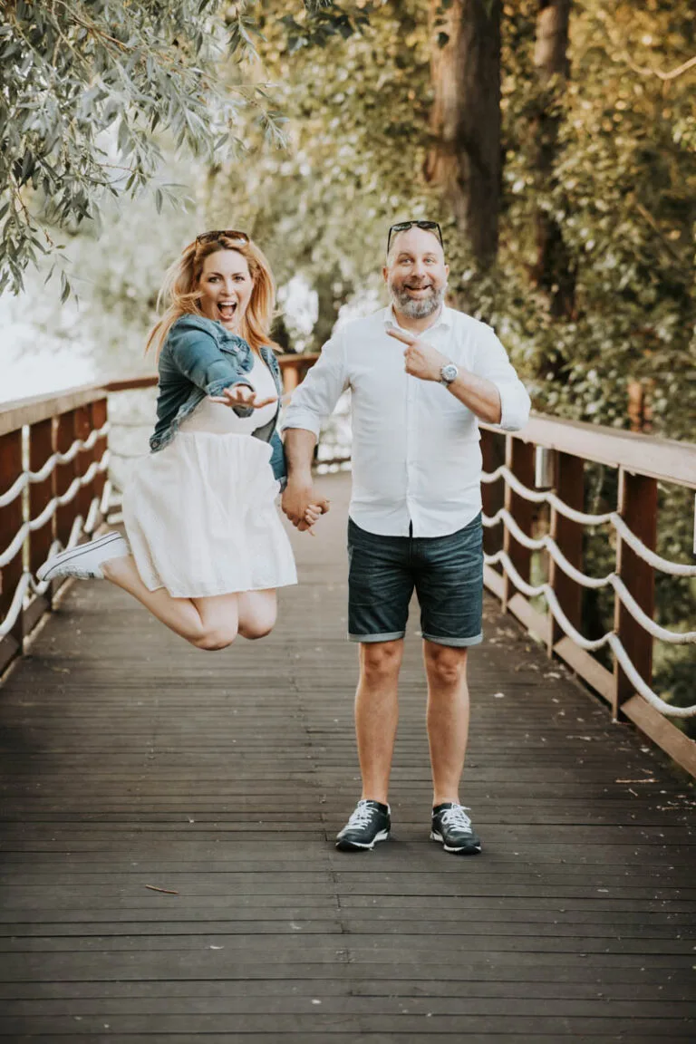 Pár egy fából készült hídon, ahol a nő ugrik örömében, fehér ruhában és farmerdzsekiben, míg a férfi fehér ingben és rövidnadrágban áll mellette, mosolyogva, rámutatva. A háttérben zöldellő fák láthatók.