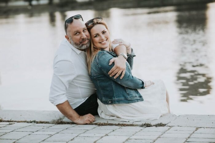 Egy szerelmes pár ül a vízparti köveken; a nő farmerdzsekiben és fehér ruhában mosolyog, miközben a férfi fehér ingben öleli át. A háttérben a víz tükröződése adja a romantikus hangulatot.