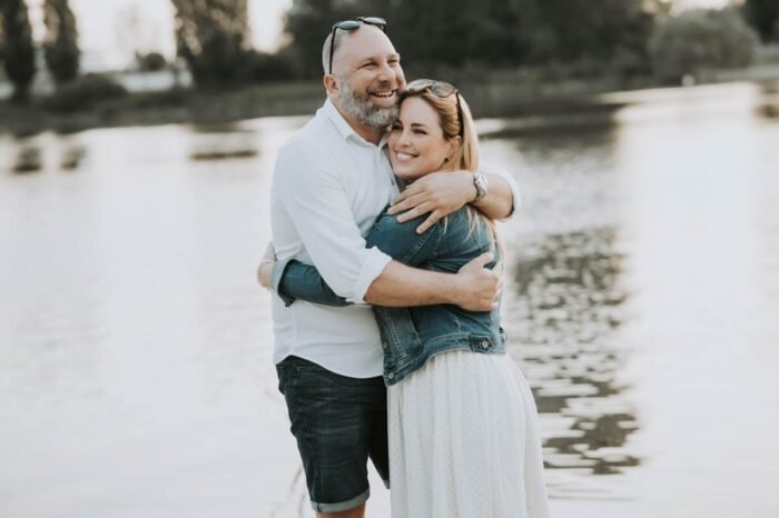 Egy mosolygós pár öleli egymást a vízparton állva; a nő farmerdzsekiben és fehér ruhában, a férfi fehér ingben és rövidnadrágban látható. A háttérben nyugodt víz és zöldellő fák adják a meghitt hangulatot.