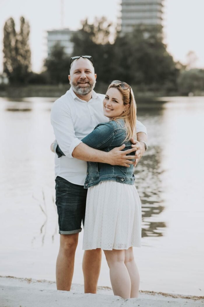 Egy boldog pár ölelkezik a vízparton, a nő fehér ruhában és farmerdzsekiben, a férfi fehér ingben és rövidnadrágban. A háttérben zöld fák és épületek tükröződnek a víz felszínén.