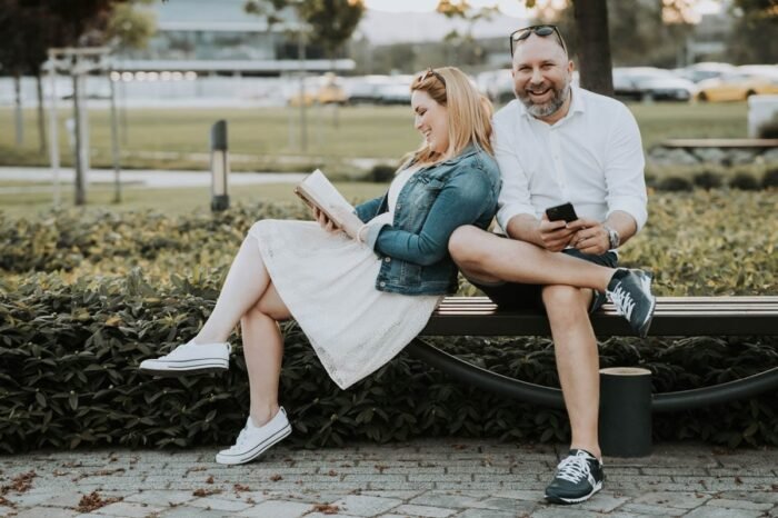 Egy pár egy parkban ül a padon; a nő könyvet olvas, miközben mosolyog, a férfi pedig telefonját nézi vidáman. A háttérben zöld növények és autók láthatók, ami modern városi környezetet idéz.