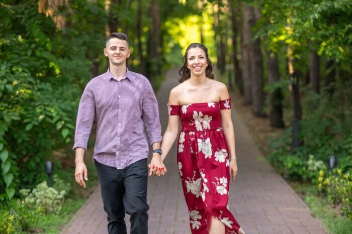 A pár kézen fogva sétál egy gyönyörű, fás sétaúton, miközben mindketten mosolyognak. A nő piros virágmintás ruhát visel, a férfi kockás inget és sötét nadrágot. A háttérben zöld fák és kellemes napfény látható, amely romantikus hangulatot kölcsönöz a pillanatnak.