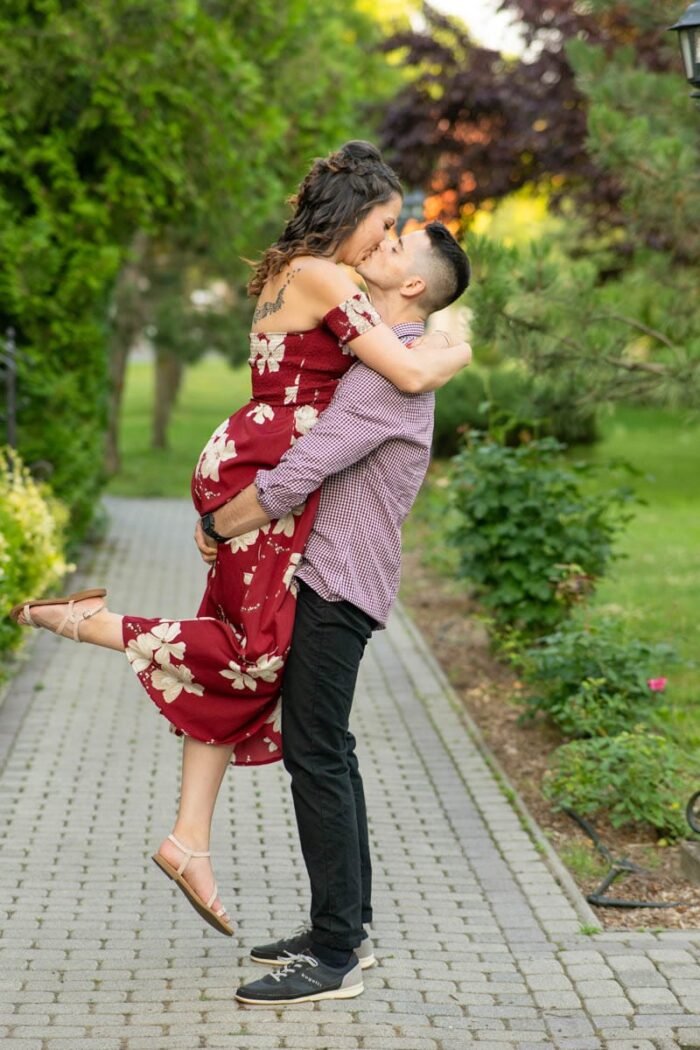 A pár sétál a parkban, és a férfi felemeli a nőt, miközben az arcuk egy pillanatra összeér. Mindketten boldogan mosolyognak, miközben a nő a férfi nyakába kapaszkodik, a férfi pedig a derekát tartja. A háttérben zöld növények és sétányok láthatók, a nő piros, virágmintás ruhát visel, a férfi pedig kockás inget és sportcipőt.