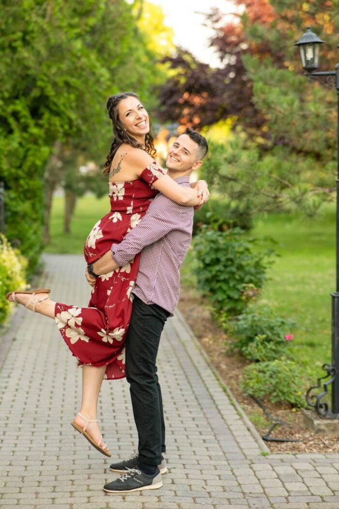 A pár boldogan nevetve, mosolygósan öleli egymást egy parkban. A férfi felemeli a nőt, miközben ő kinyújtja a lábát és boldogan nevet. A háttérben zöld növények és egy jól karbantartott sétány látható, amely romantikus hangulatot ad a fotóhoz. A nő virágmintás piros ruhát visel, a férfi pedig kockás inget, mindketten sportcipőt viselnek.