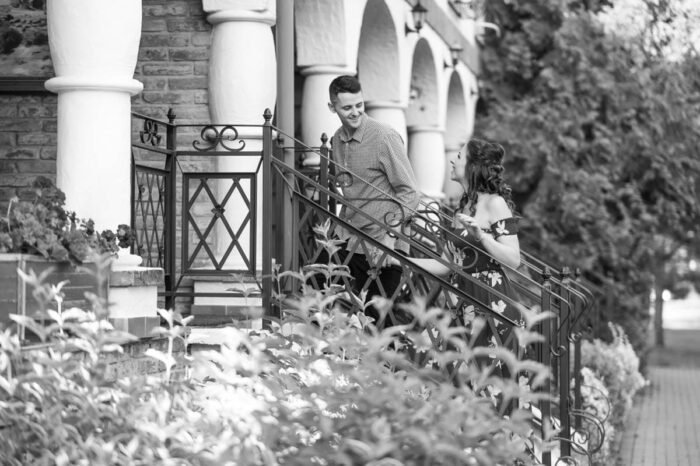 Egy fiatal jegyespár boldog pillanatait örökíti meg a fotó, ahol a férfi és nő egy kovácsoltvas korláton állva mosolyognak egymásra. A háttérben látható egy téglaépület, amely klasszikus, elegáns hangulatot ad a képnek. A nő virágmintás, strapabíró ruhát visel, a férfi kockás inget és sötét nadrágot. A környező zöld növényzet kellemesen lágyítja az összhatást, miközben az erkély és a lépcső adja meg a romantikus helyszínt.
