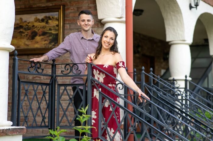 Egy mosolygó fiatal jegyespár áll egy kovácsoltvas korlátos erkélyen. A háttérben téglafal és egy faajtó látható, amelyen "Árbo lakosztály" felirat olvasható. A férfi kockás inget visel, míg a nő virágmintás bordó ruhát, amely elegáns és romantikus hangulatot sugároz.