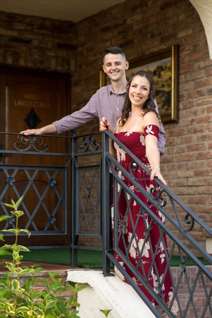 Egy mosolygó fiatal jegyespár áll egy kovácsoltvas korlátos erkélyen. A háttérben téglafal és egy faajtó látható, amelyen "Árbo lakosztály" felirat olvasható. A férfi kockás inget visel, míg a nő virágmintás bordó ruhát, amely elegáns és romantikus hangulatot sugároz. A zöld növényzet az előtérben természetes keretet ad a képnek, amely barátságos és meghitt pillanatot örökít meg.