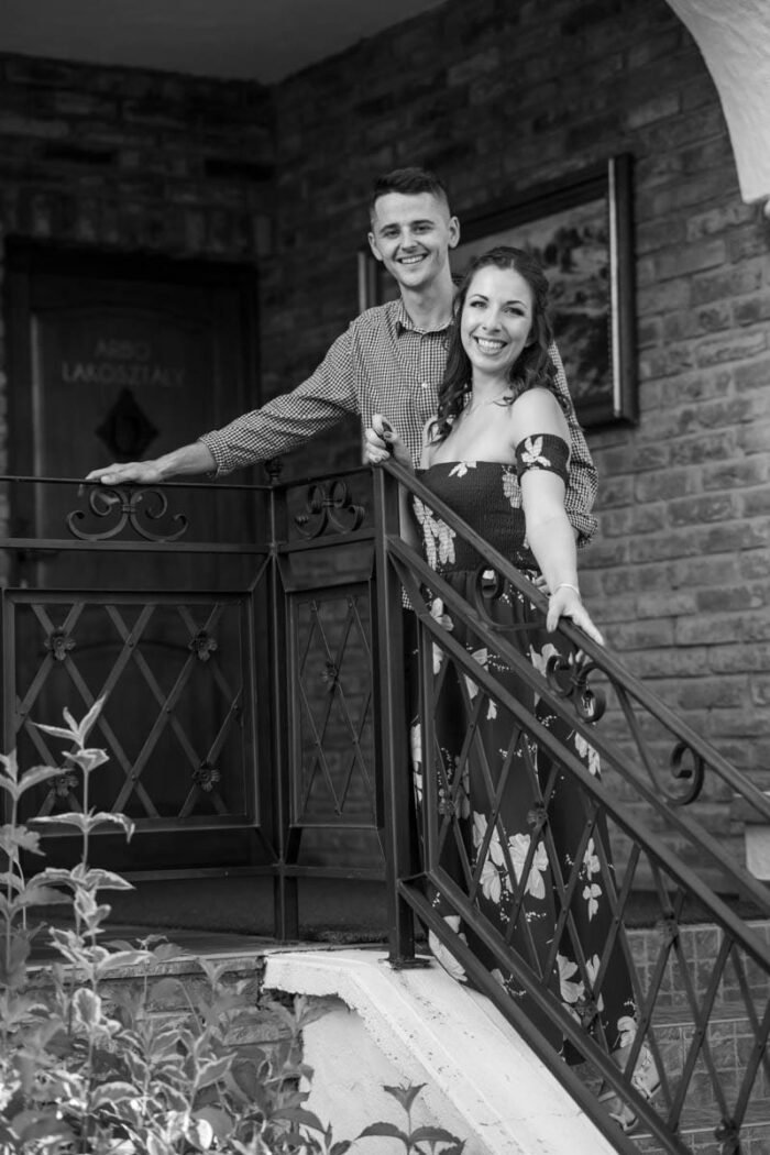 Egy fiatal jegyespár mosolyog a kamerába, miközben egy kovácsoltvas korláttal díszített erkélyen állnak. A háttérben téglafal, faajtó és egy festmény látható, amelyek meleg, otthonos hangulatot kölcsönöznek a képnek. A fekete-fehér stílus kiemeli az érzelmeket és az időtlenséget. A férfi kockás inget, a nő pedig virágmintás ruhát visel, miközben gyengéd közelségben állnak egymás mellett. Egy fiatal jegyespár mosolyog a kamerába, miközben egy kovácsoltvas korláttal díszített erkélyen állnak. A háttérben téglafal, faajtó és egy festmény látható, amelyek meleg, otthonos hangulatot kölcsönöznek a képnek. A fekete-fehér stílus kiemeli az érzelmeket és az időtlenséget. A férfi kockás inget, a nő pedig virágmintás ruhát visel, miközben gyengéd közelségben állnak egymás mellett.