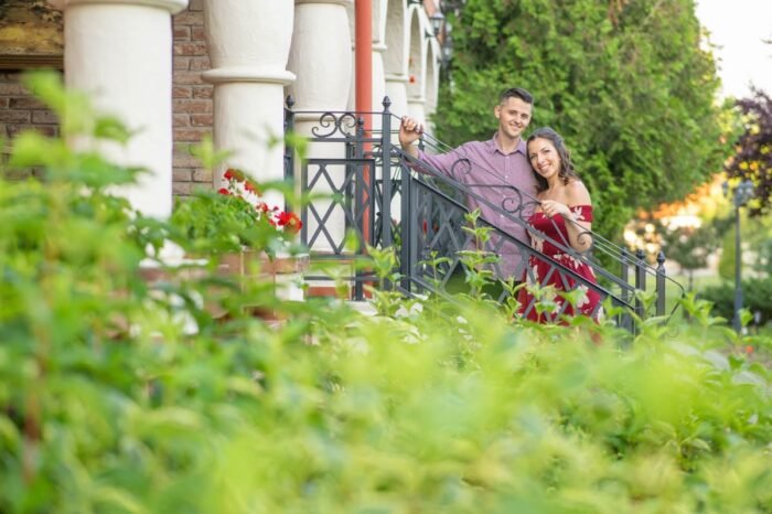 Egy mosolygós jegyespár áll a lépcső korlátjánál, körülöttük buja zöld növényzet és élénk színű virágok díszítik a jelenetet. A férfi kockás inget visel, miközben szeretetteljesen néz párjára, aki piros, virágmintás ruhát hord. A háttérben egy oszlopos veranda és tégla falak fokozzák a romantikus hangulatot.