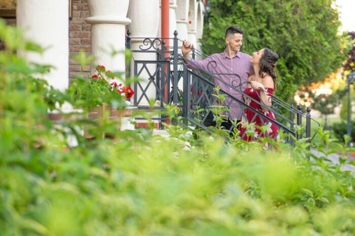 Egy jegyespár romantikus jelenete egy virágokkal és növényekkel körülvett lépcsőn. A férfi kockás inget visel, és a korlátra támaszkodik, miközben mosolyogva néz a piros, virágos ruhát viselő nőre, aki szeretetteljesen tekint rá. A háttérben oszlopok és buja növényzet látható, ami a jelenet meghitt hangulatát fokozza.