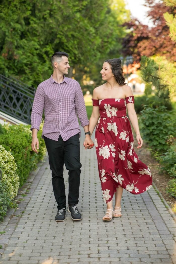 Egy mosolygó jegyespár kézen fogva sétál egy parkosított ösvényen. A férfi kockás ingben és fekete nadrágban van, a nő piros, virágmintás ruhát visel, és egymásra néznek szeretetteljes pillantással. A háttérben zöld növények és fák láthatók, a jelenet meleg napfényben úszik.